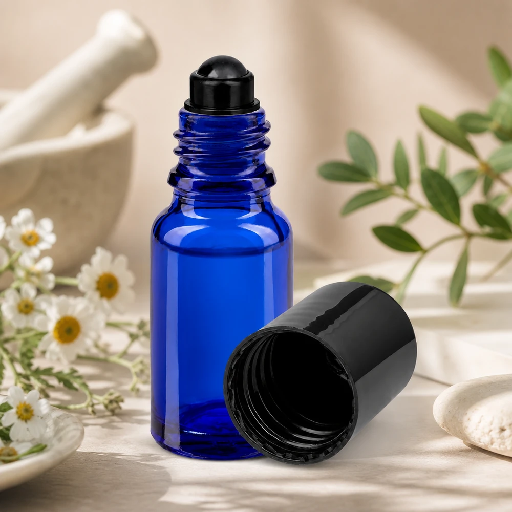 Butelka apteczna niebieska 10ml zakrętka roll-on czarna Niebieska buteleczka typu roll-on o pojemności 5 ml, model STD, z czarną nakrętką, obok ziół w moździerzu i kwiatów rumianku