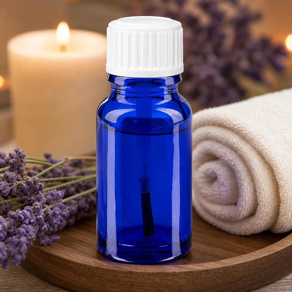 Butelka apteczna niebieska 10ml zakrętka biały pędzelek Buteleczka do aromaterapii o pojemności 10 ml, wykonana z niebieskiego szkła aptecznego, z białą nasadką z pędzelkiem, obok kwiatów lawendy