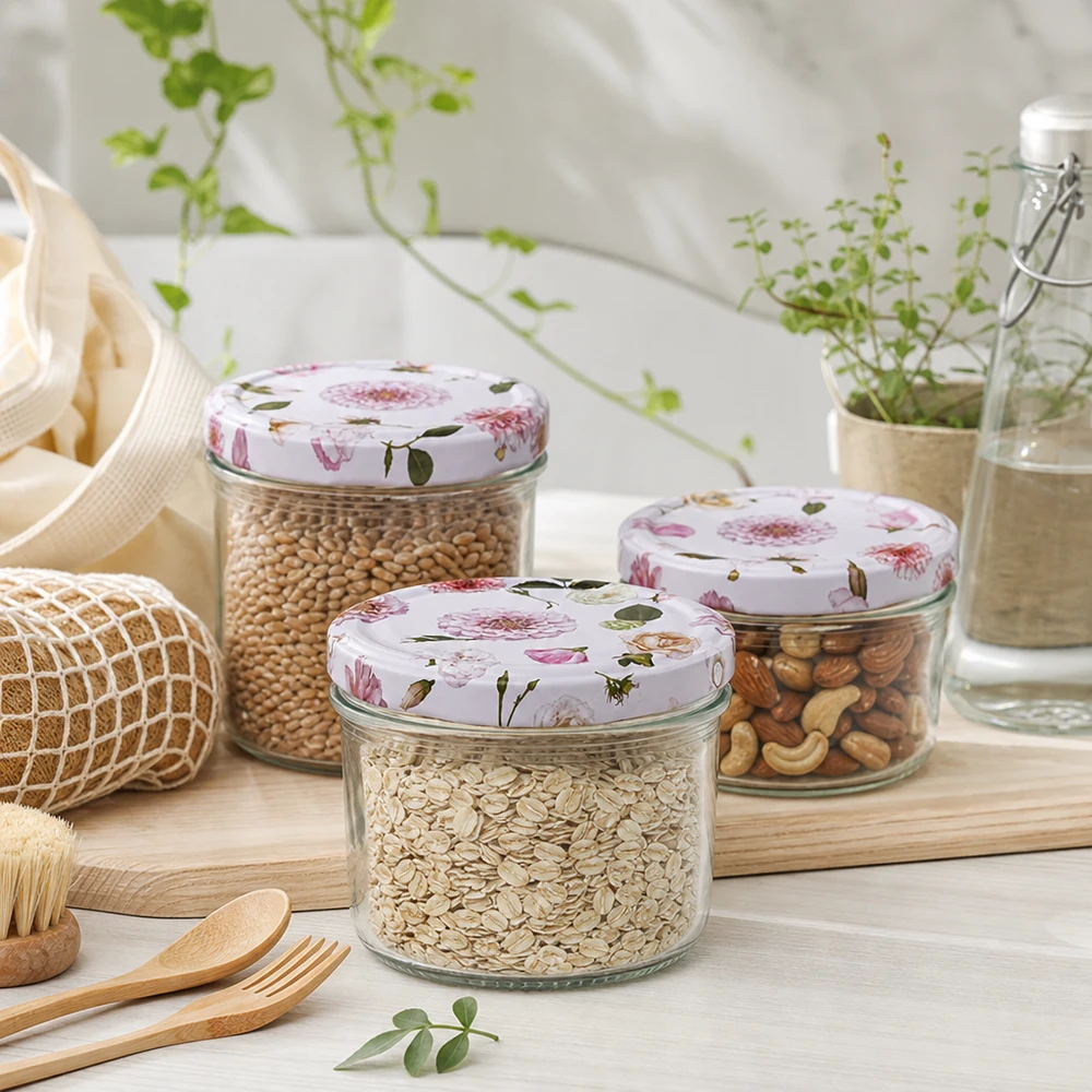 Trzy słoiki cylindryczne o pojemności 230 ml z różowymi pokrywkami w kwiaty wypełnione zbożami i orzechami w zrównoważonym środowisku kuchennym