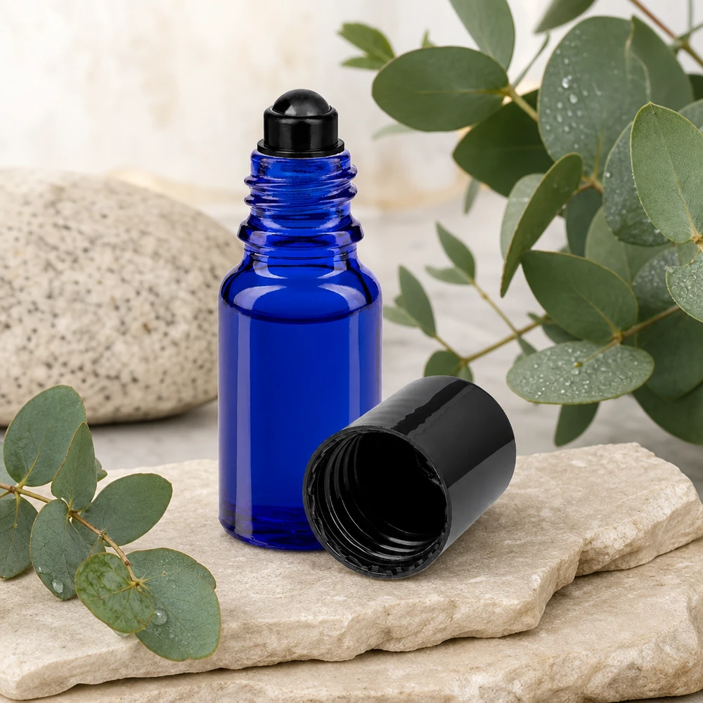 Butelka apteczna niebieska 10ml zakrętka roll-on czarna Niebieska buteleczka ApoGlas z kulką o pojemności 10 ml z czarną nakrętką, stojąca obok gałązek eukaliptusa na naturalnym kamieniu