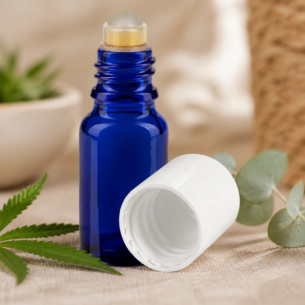 Butelka apteczna niebieska 10ml nakrętka roll-on biała Niebieska buteleczka z kulką o pojemności 10 ml z białą nakrętką, umieszczona obok liści konopi i eukaliptusa na płótnie lnianym