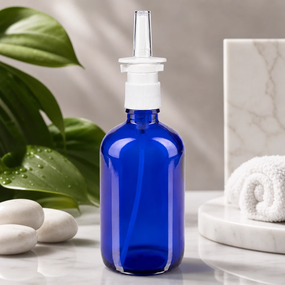 Szklana butelka o pojemności 100 ml w kolorze kobaltowym z nasadką do rozpylania do nosa, stojąca na białym marmurze obok kamieni do aromaterapii