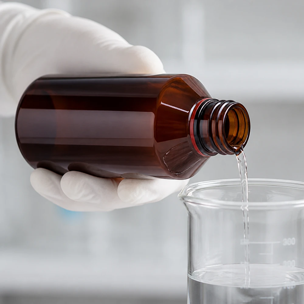 Osoba w rękawiczkach przelewa przezroczysty płyn z bursztynowej butelki PET o pojemności 200 ml do szklanej zlewki w laboratorium