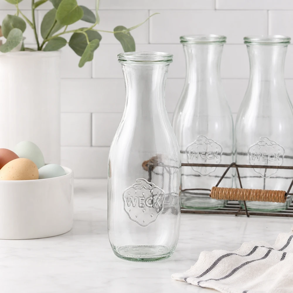 Butelka na sok o pojemności 1062 ml z szklaną pokrywką WECK RR60 Butelka na sok WECK 1062 ml pusta szklana butelka w nowoczesnej białej kuchni z drucianym koszem na jajka i rośliną