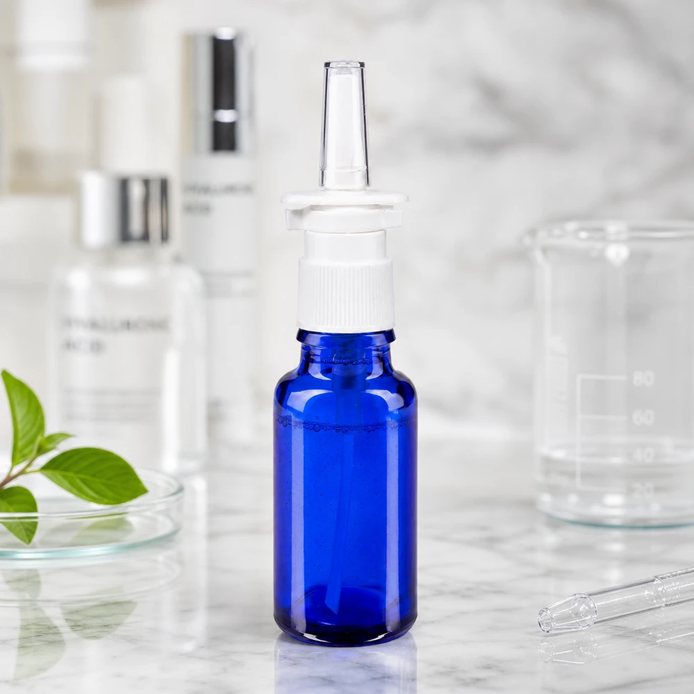 Butelka z aerozolem do nosa o pojemności 20 ml, wykonana z niebieskiego szkła aptecznego, z białym rozpylaczem, obok szklanej zlewki i kwasu hialuronowego