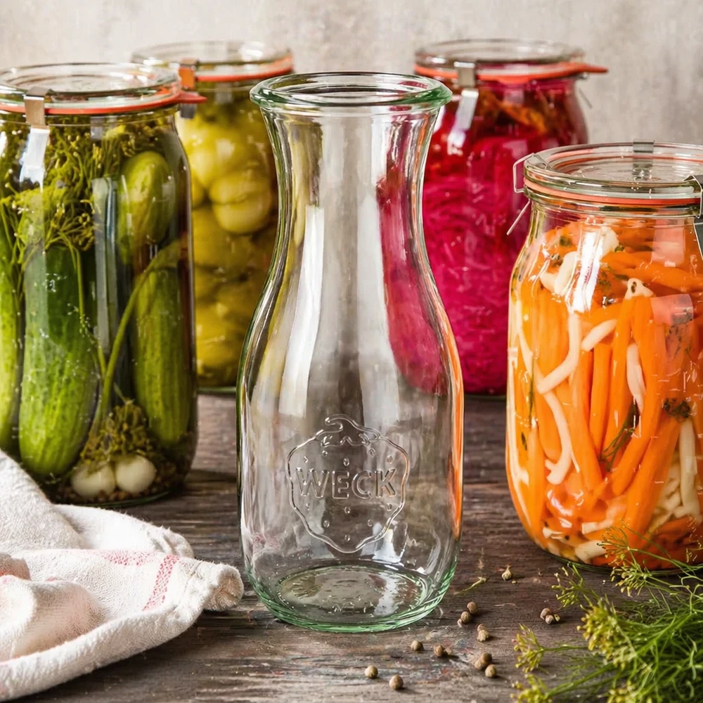 Butelka na sok WECK RR60 o pojemności 530 ml Butelka na sok WECK RR60 o pojemności 530 ml otoczona kolorowymi słoikami z kiszonymi warzywami i akcesoriami do przetworów na drewnianym tle