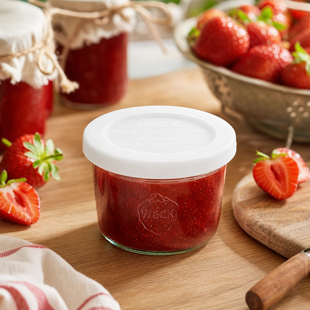 Słoik o pojemności 80 ml z pokrywką zachowującą świeżość WECK RR60 Kubki WECK 80 ml RR60 wypełnione dżemem truskawkowym na drewnianej desce w otoczeniu świeżych truskawek i słoików z dżemem