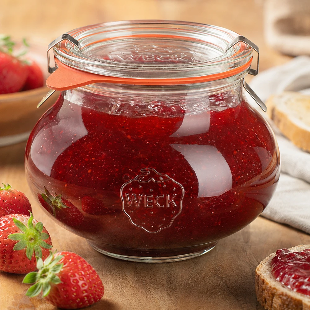 Kompletny słoik dekoracyjny WECK RR80 o pojemności 560 ml Dekoracyjny słoik WECK 560ml wypełniony domowym dżemem truskawkowym, zamknięty uchylną pokrywką, otoczony świeżymi owocami i warzywami.