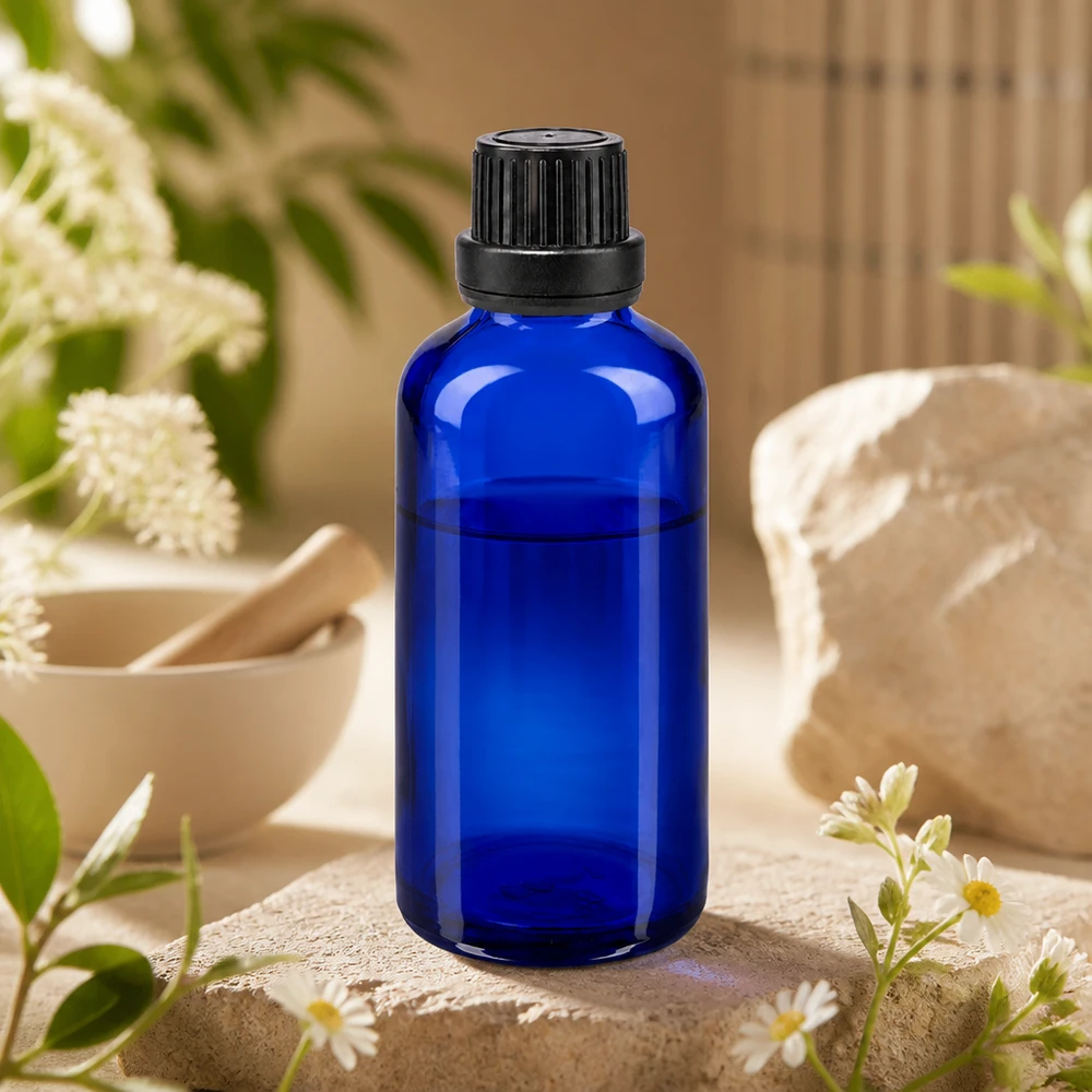 Szklana butelka o pojemności 50 ml w kolorze kobaltowym z czarną zakrętką, stojąca na kamieniu naturalnym z kwiatami rumianku i moździerzem
