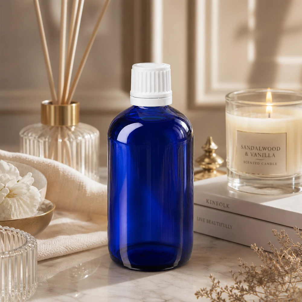 Butelka apteczna niebieska 100ml zakrętka biała OV Niebieska szklana buteleczka apteczna o pojemności 100 ml z białą nakrętką zabezpieczoną przed dziećmi, stojąca na marmurze obok świecy o zapachu drzewa sandałowego