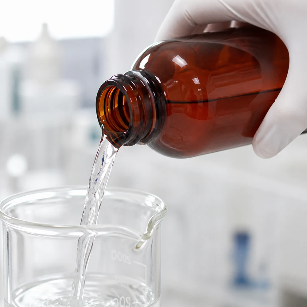 Butelka PET 250 ml z niebieską nakrętką OV Laborant, mając na rękach rękawiczki ochronne, przelewa przezroczysty płyn z brązowej butelki PET o pojemności 250 ml do zlewki.