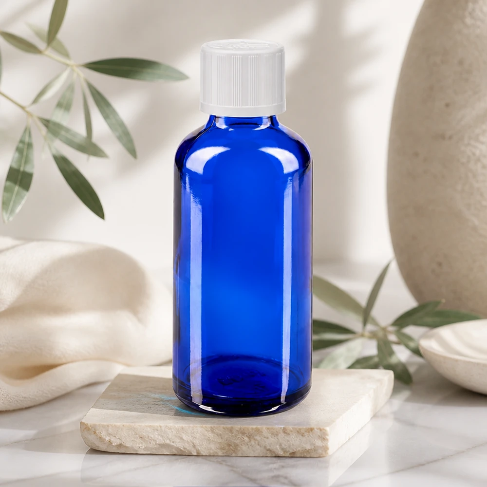 Niebieska szklana butelka o pojemności 50 ml z białą zakrętką, stojąca na marmurowej płycie w jasnym, naturalnym otoczeniu